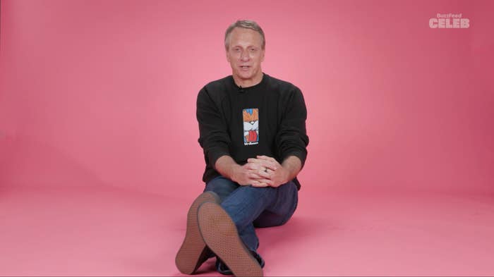 Man sitting with legs crossed, wearing a casual black sweatshirt with a graphic, on a pink background