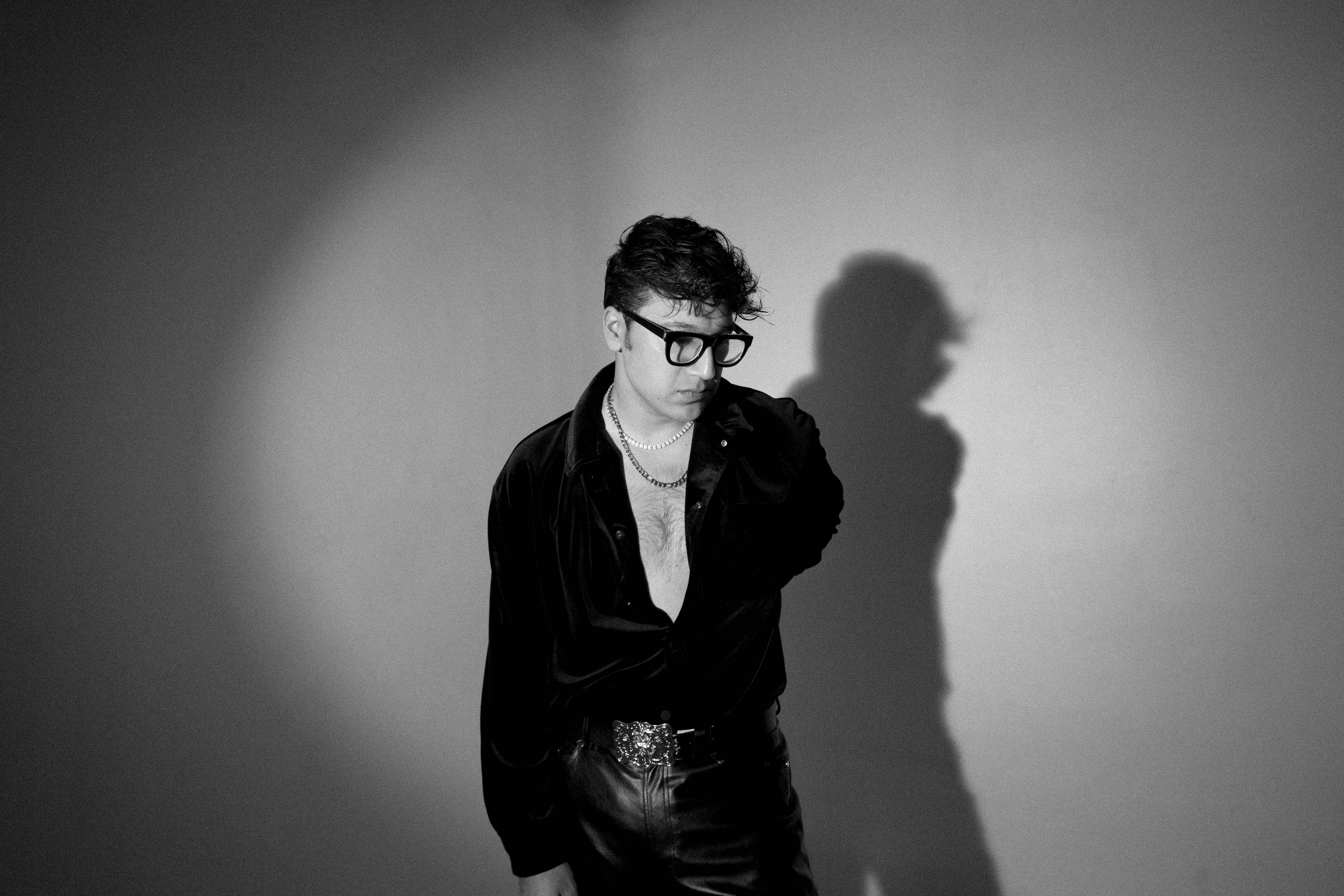 Persona con camisa y pantalones de estilo elegante, usando gafas. Está de pie con una mano en el bolsillo, proyectando una sombra en la pared