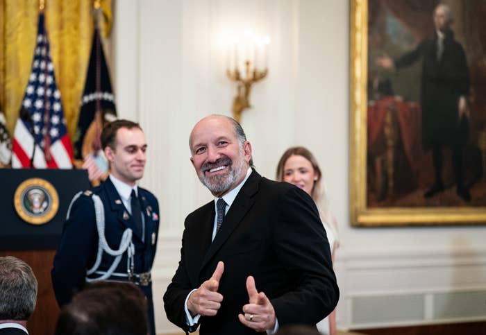 A smiling man in a suit gives two thumbs up at a formal event. Flags and a portrait adorn the background