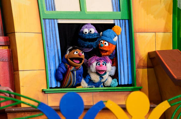 Puppet characters from a TV show, including a furry blue one and a pink one with a scarf, appear happily at a window