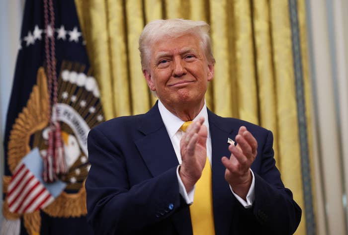 A person in a suit clapping in a formal setting, with an American flag and presidential seal visible in the background