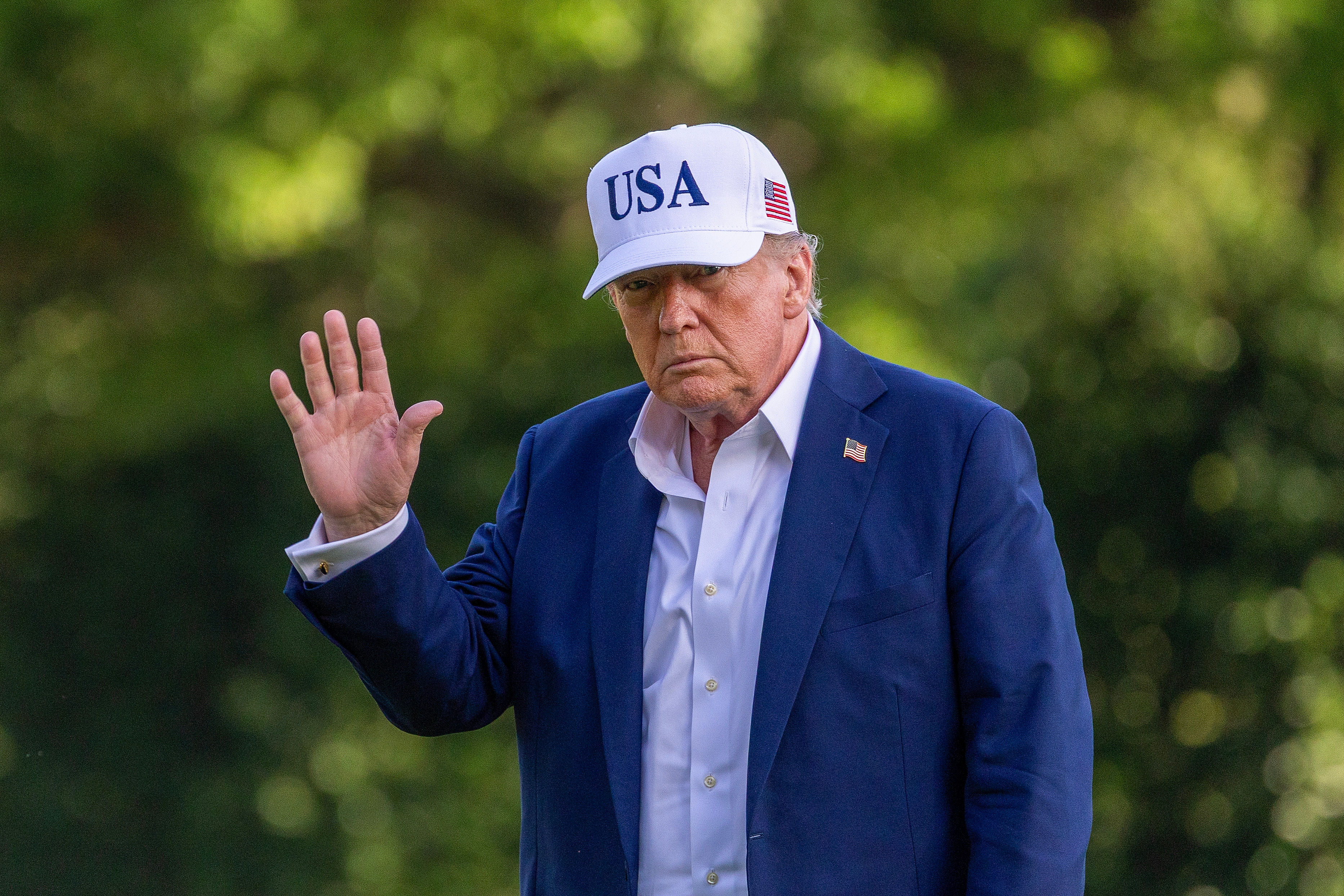 A person in a suit and white "USA" baseball cap waves outdoors in a casual setting