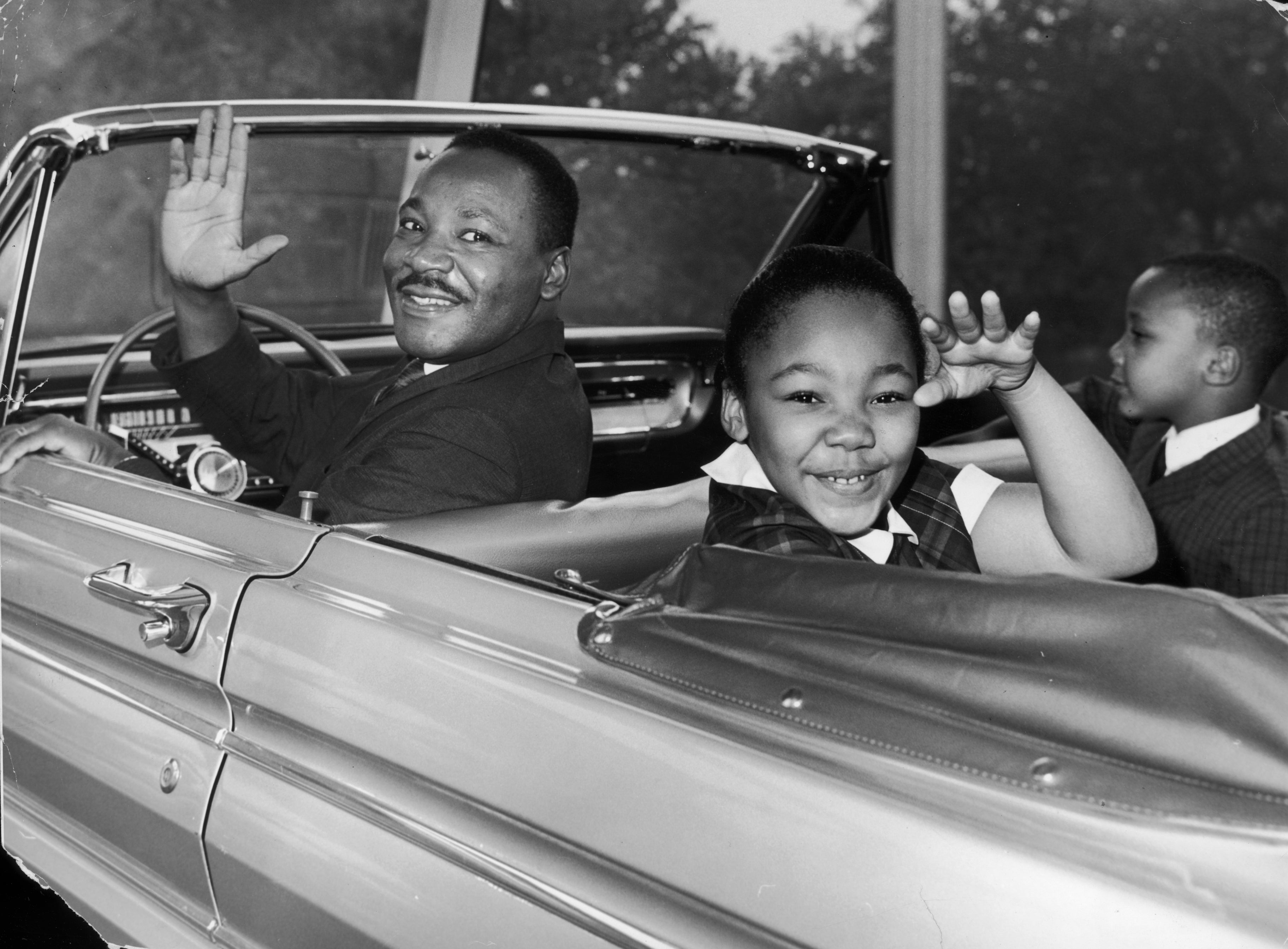 A antheral smiling and waving, seated successful a convertible, with a young kid beside him, besides waving. Another kid is seated successful the back