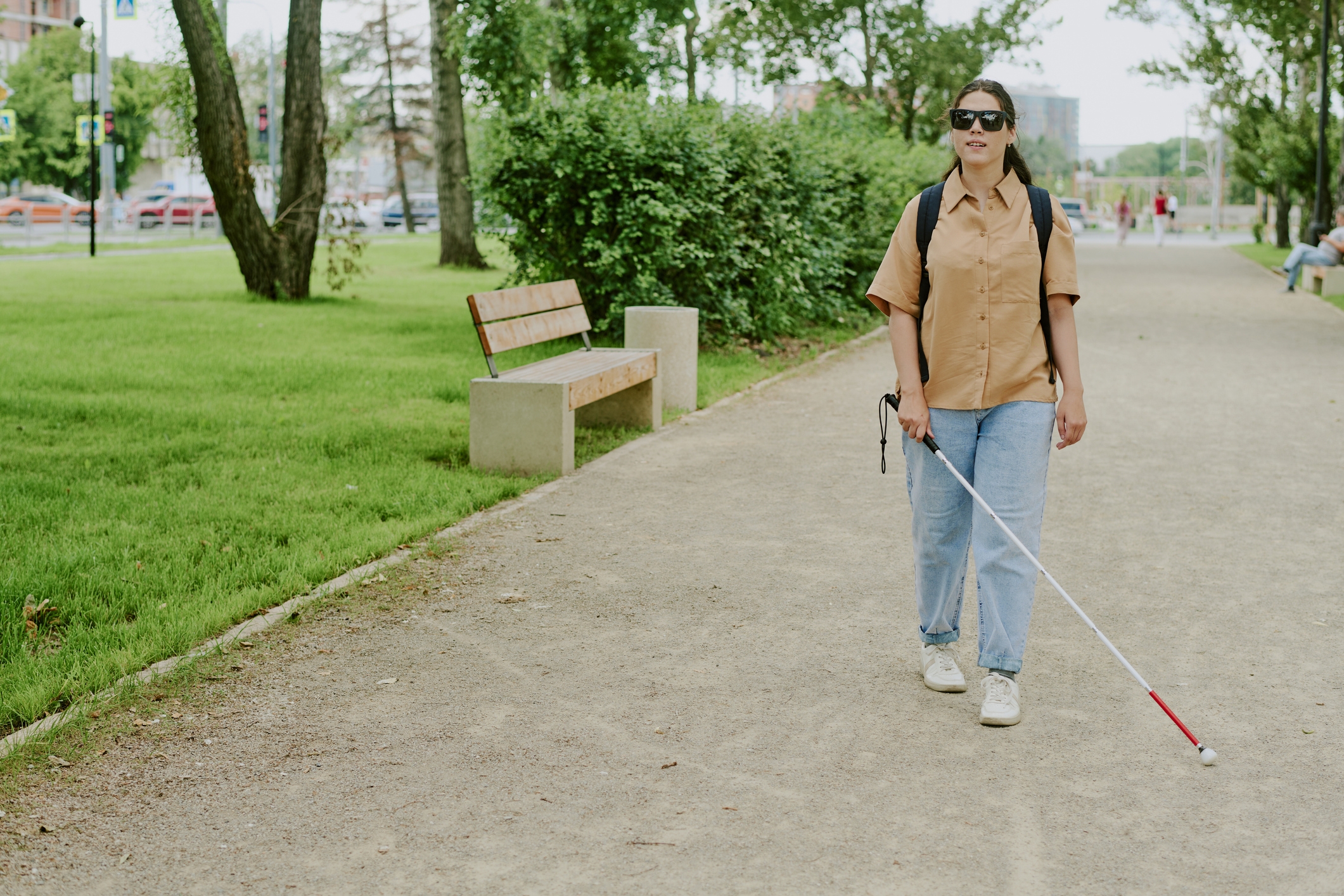 A woman with a white cane walks confidently down a park path, wearing sunglasses, a short-sleeve shirt, jeans, and a backpack