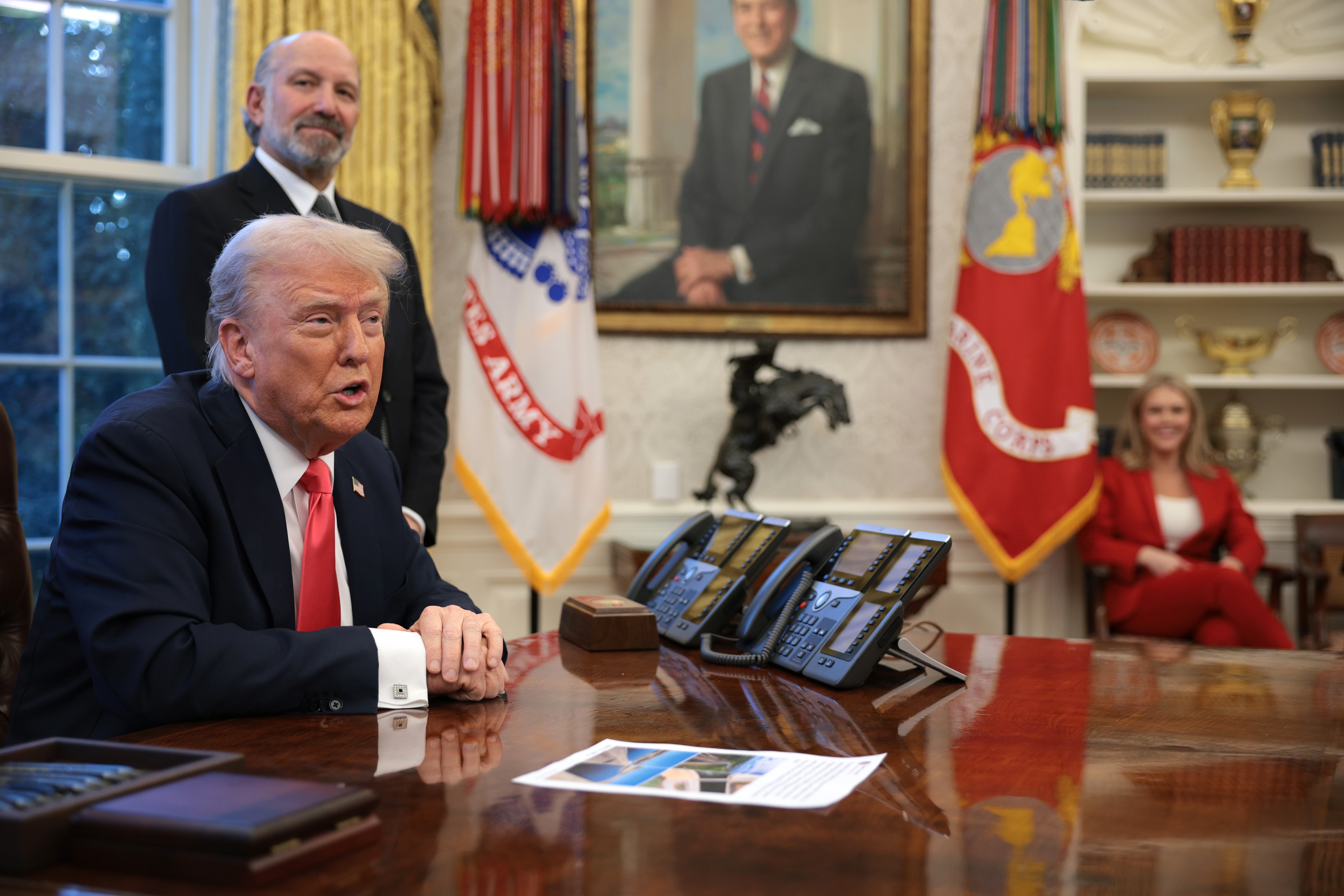 A man in a suit speaks at a desk in an official office with two individuals seated and standing nearby