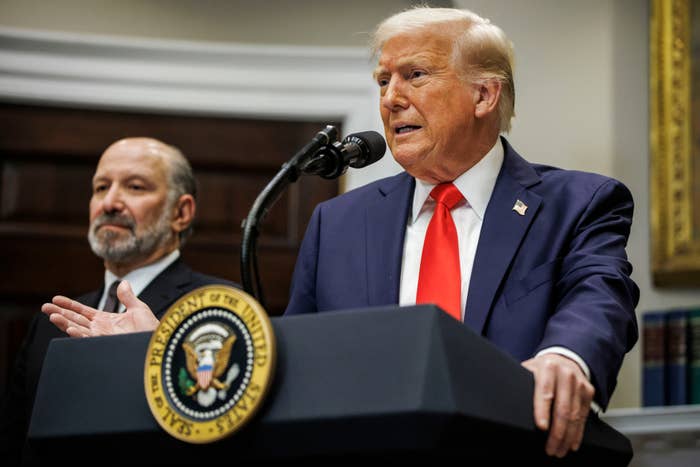 Man in suit and red tie speaking at lectern with presidential seal; another man stands in the background