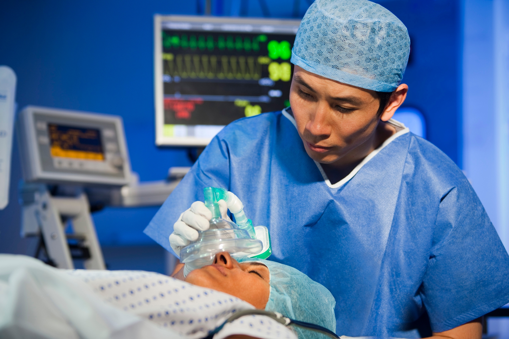 A medical professional in scrubs and a cap holds a mask over a patient's face in a hospital setting, with monitors visible in the background