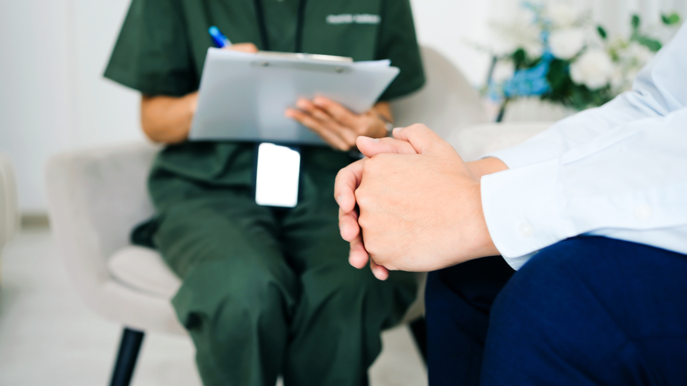 A person in a white shirt sits with clasped hands, facing a healthcare professional taking notes on a clipboard in a clinical setting