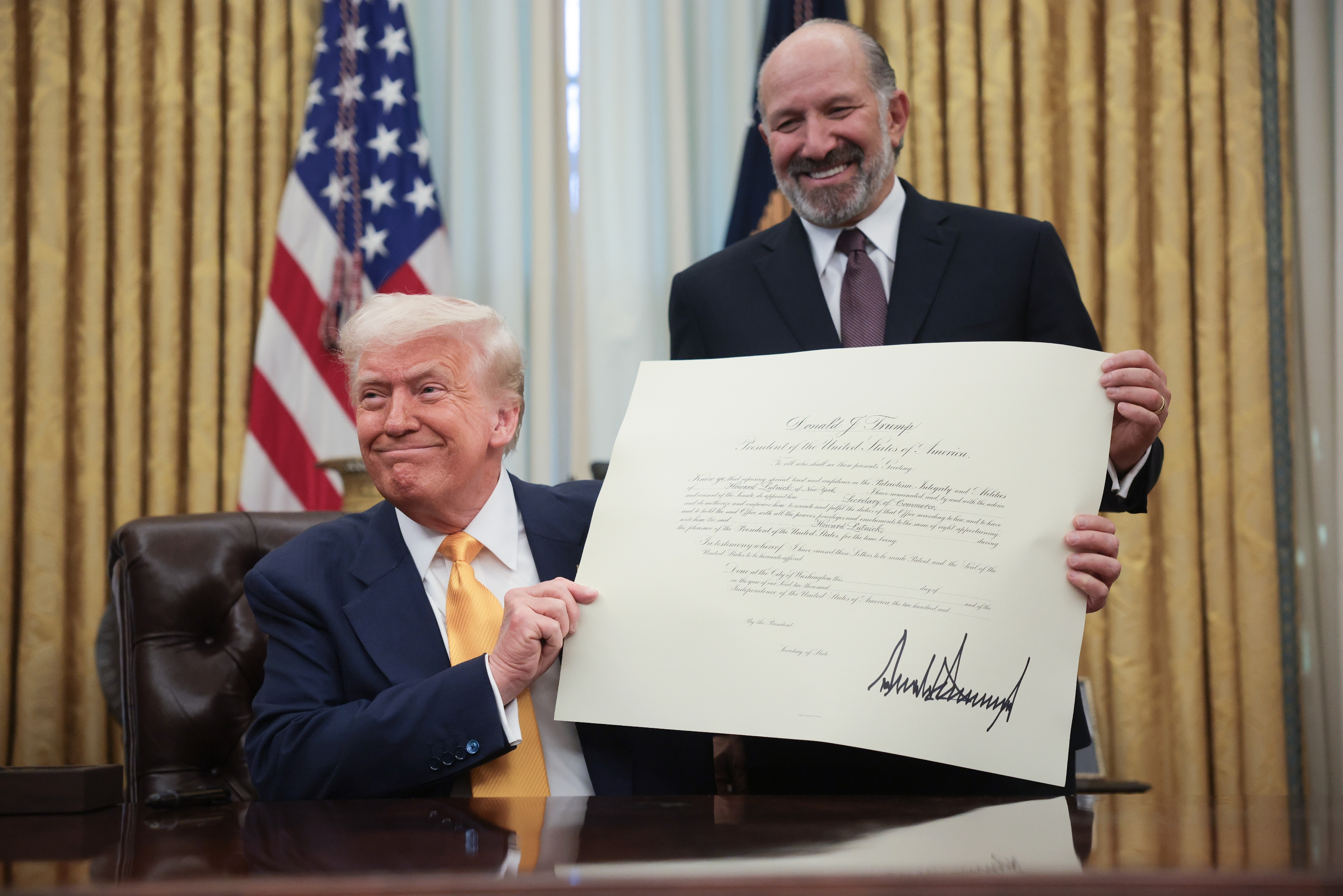 Two people in formal attire in an office. One holds a document with a signature. An American flag is in the background