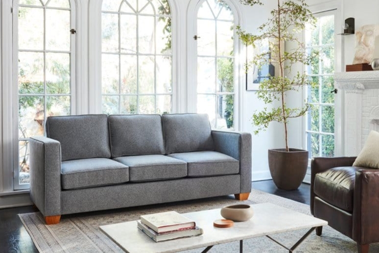 Modern living room with a gray sofa, white coffee table, and a tall indoor plant near large windows. Bright and minimalist decor