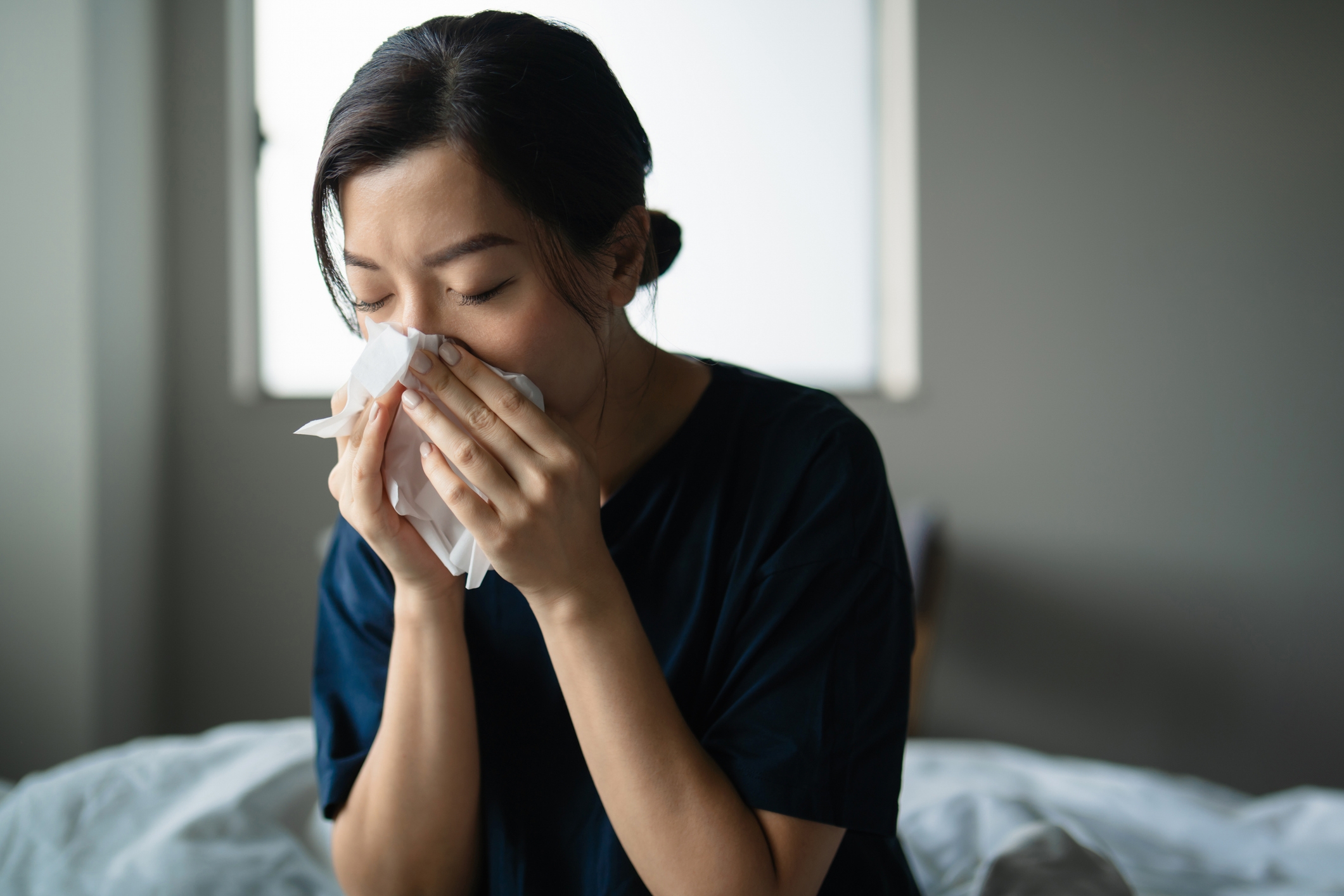 Person sitting on a bed, sneezing into a tissue, with a neutral background, capturing a moment of dealing with a cold or allergies
