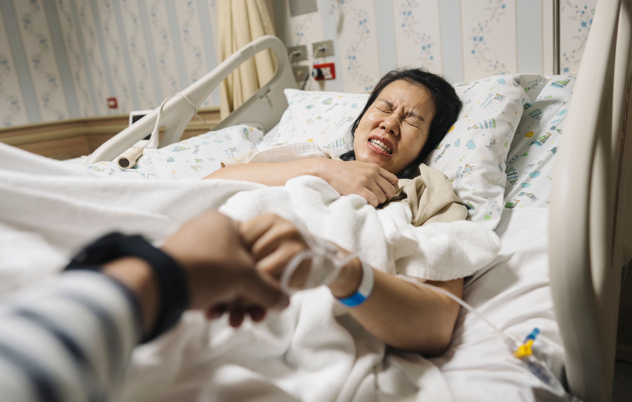 Person in hospital bed grimacing in discomfort, clutching chest. Another person holds their hand, offering support. Hospital room background