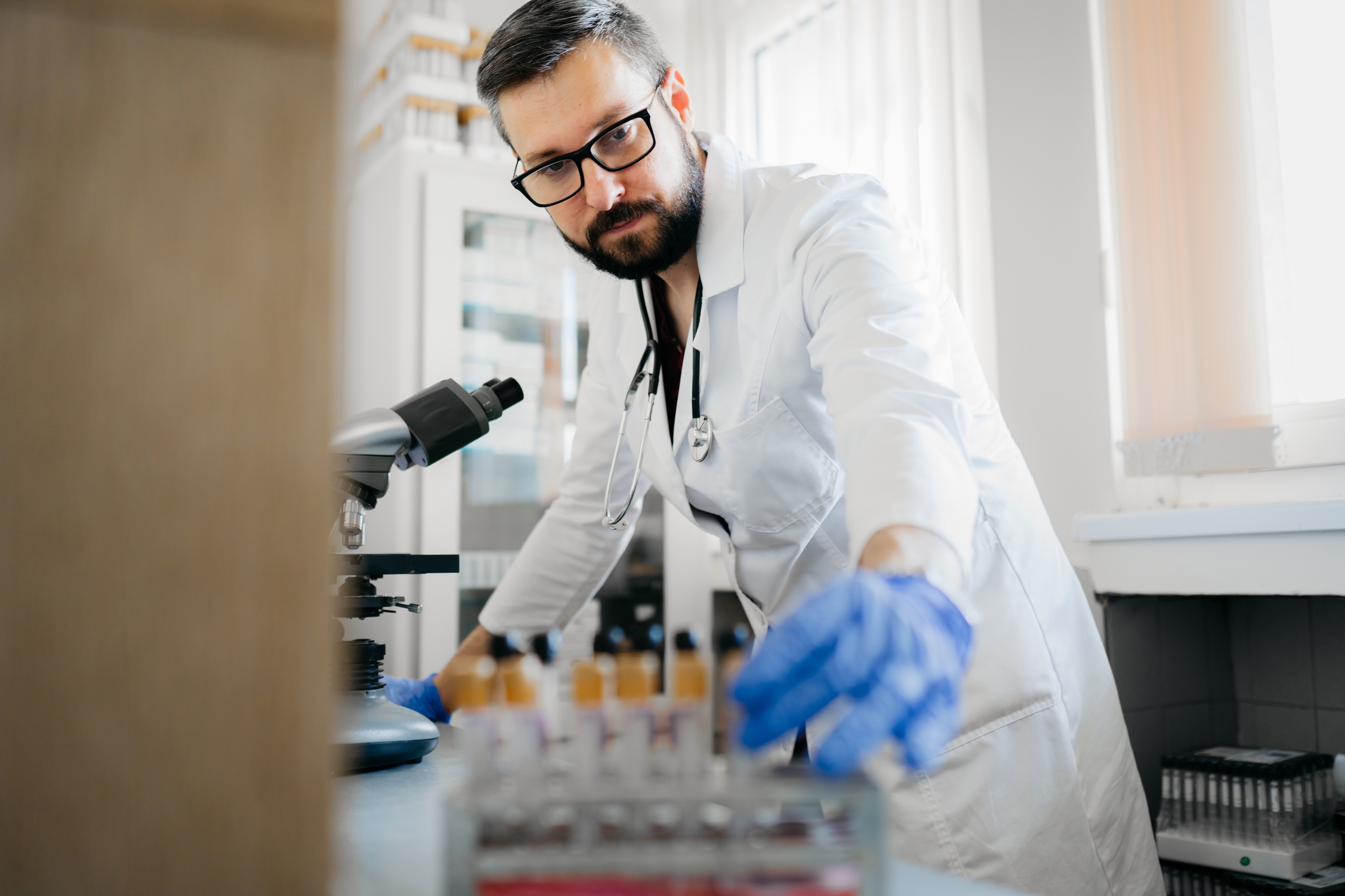 Scientist in a lab coat and gloves examines test tubes near a microscope in a laboratory setting