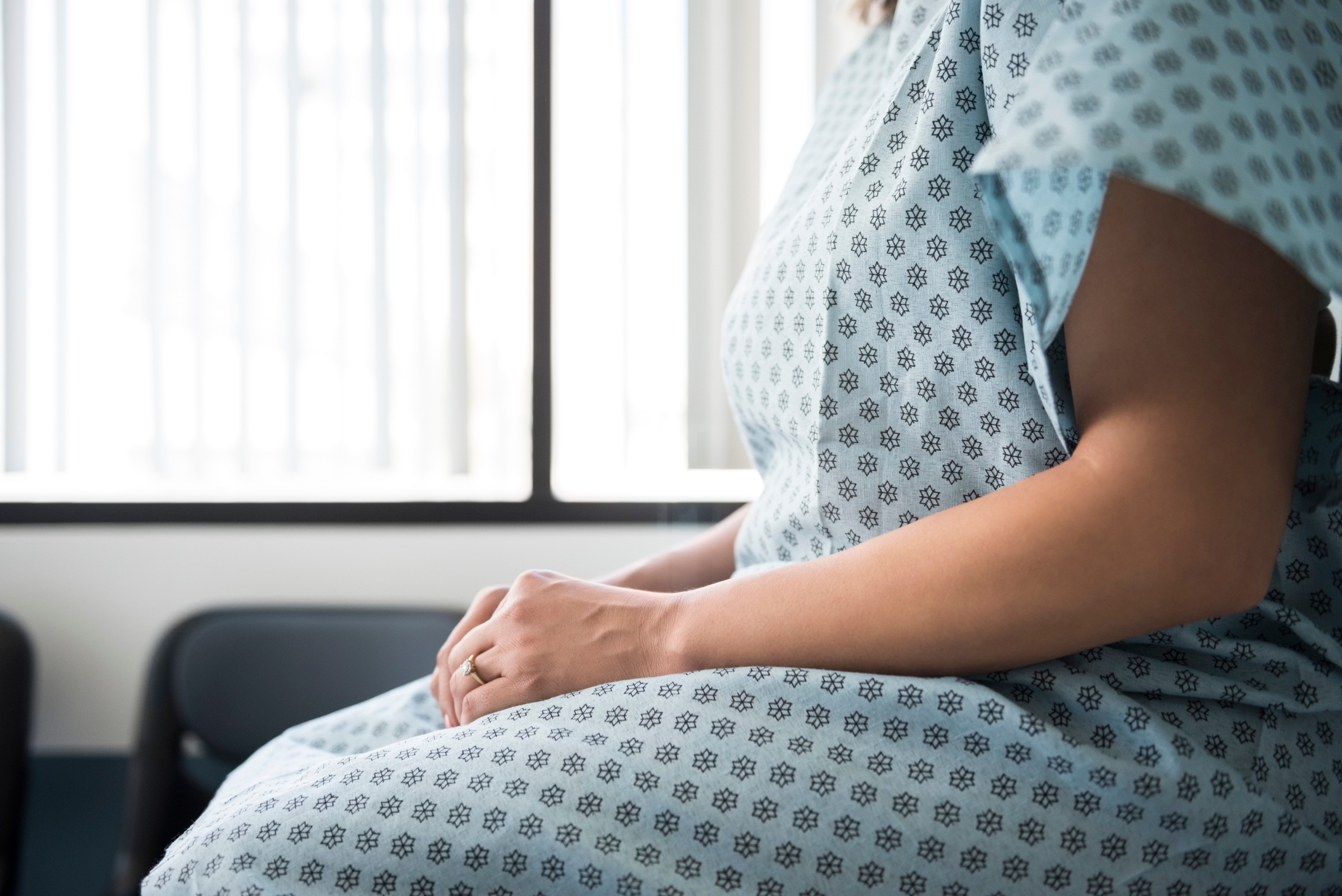 Person in a hospital gown sits in a medical exam room, partially visible window behind them