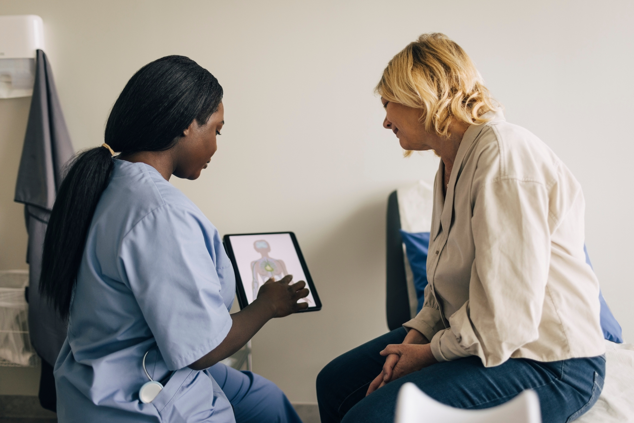 A healthcare professional in scrubs shows a patient a diagram on a tablet while sitting in a clinical setting