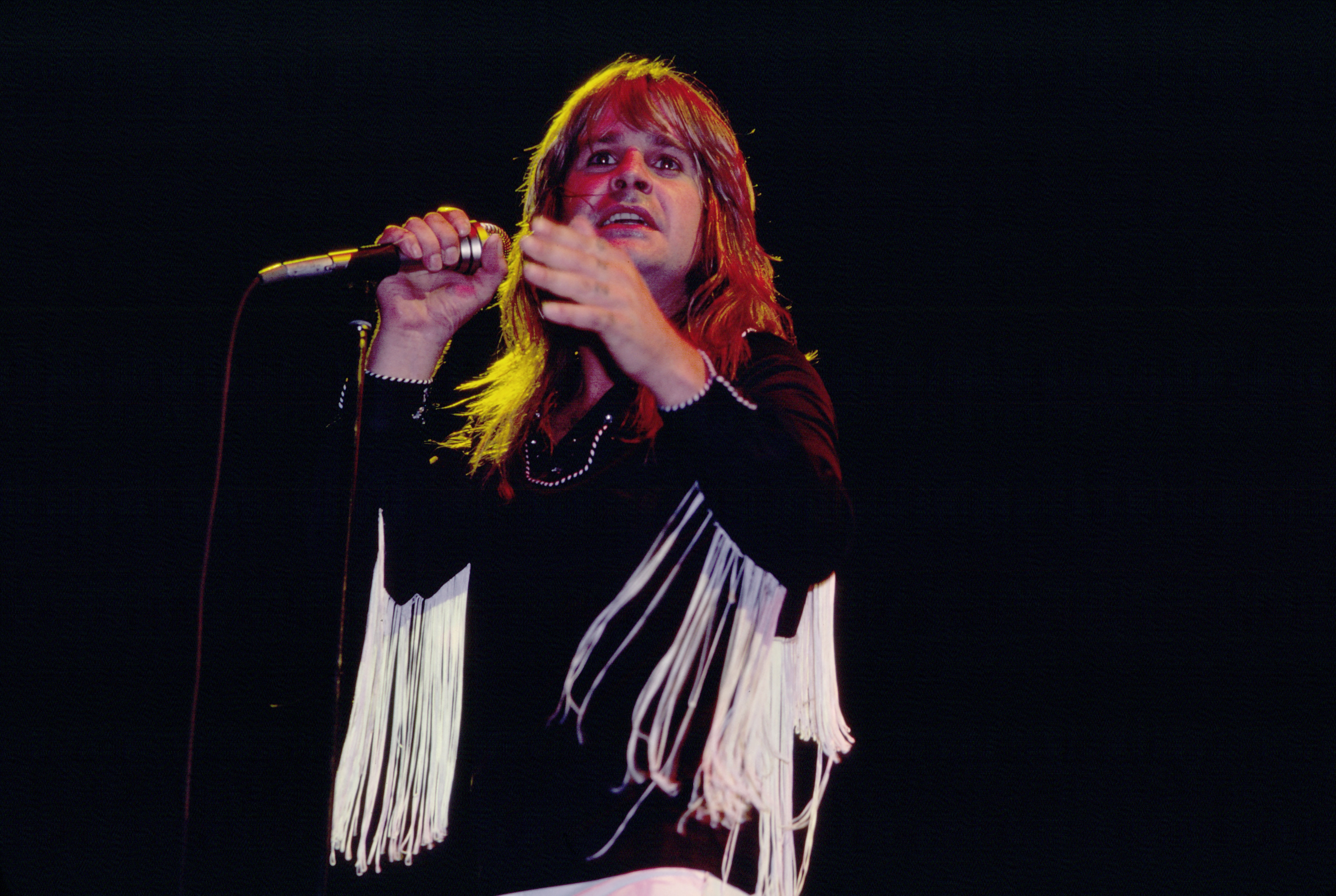 Person with long hair singing on stage, wearing a fringed jacket, holding a microphone