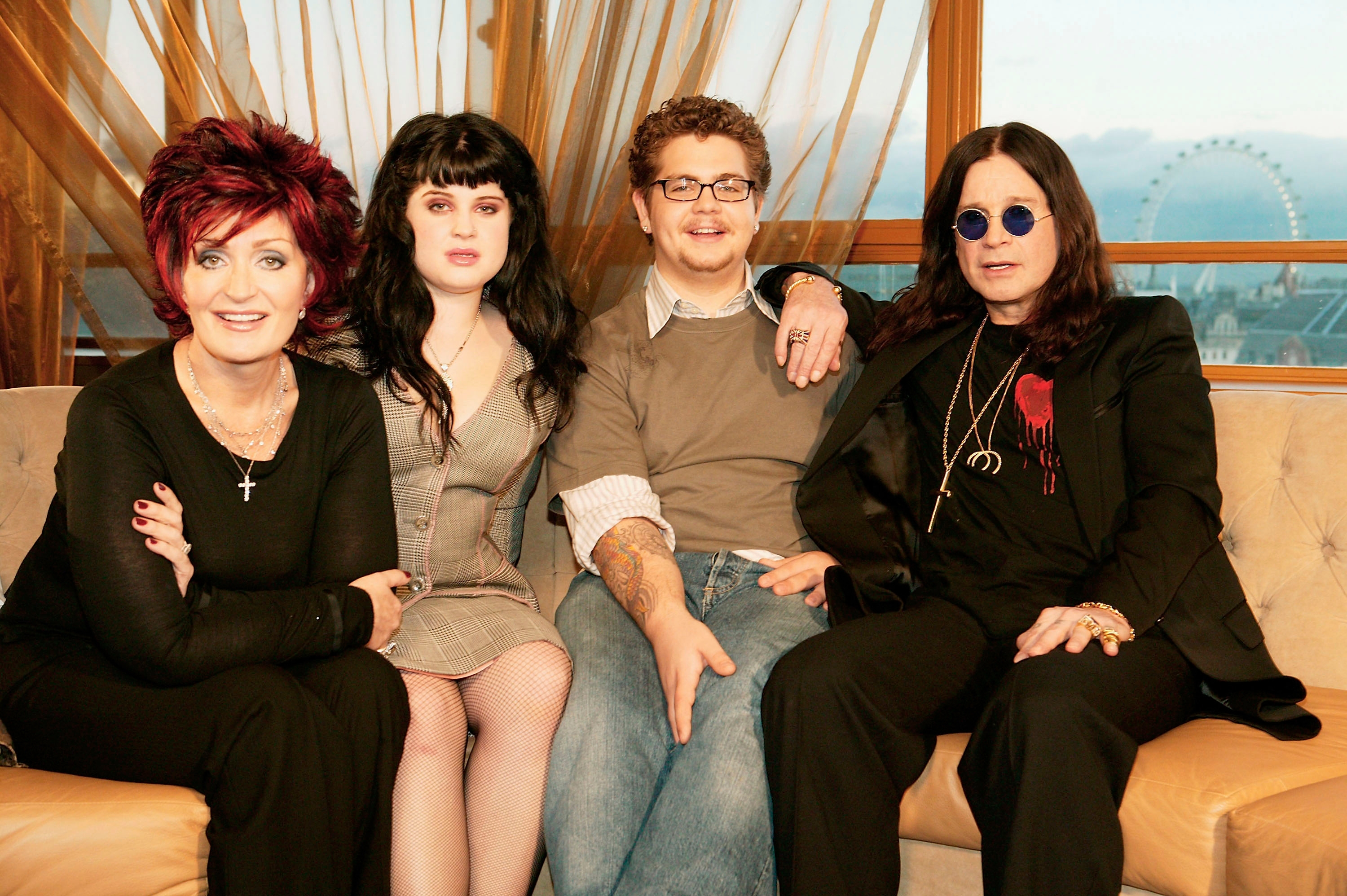Four people sit on a couch, smiling. Three wear black, where one has a red accent. Another wears a gray dress. Large window backdrop