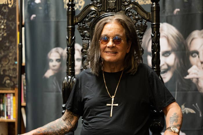 Person with long hair wearing sunglasses, a black t-shirt, and a large cross necklace, seated on an ornate chair with promotional posters behind