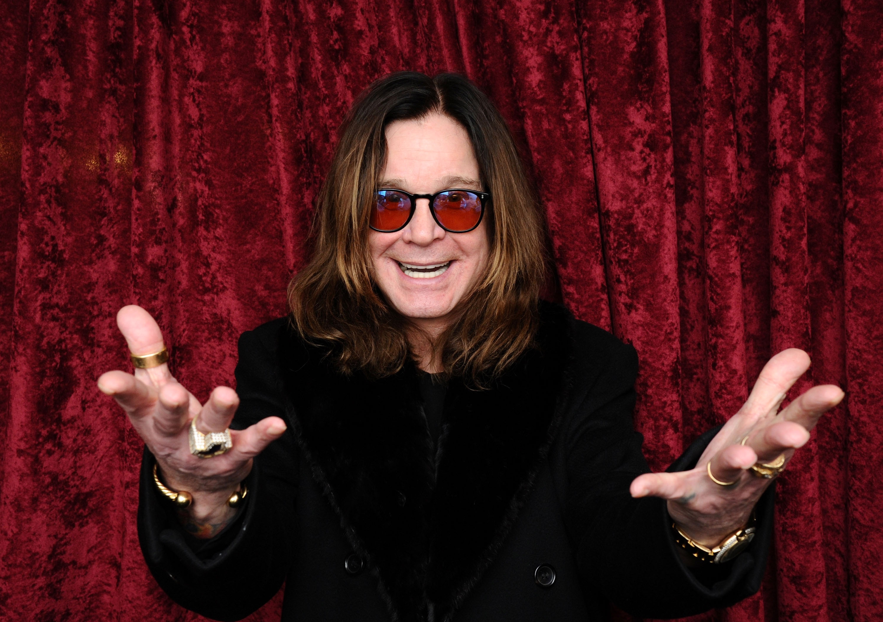 A person with long hair wearing a black coat and sunglasses poses with open hands against a textured backdrop