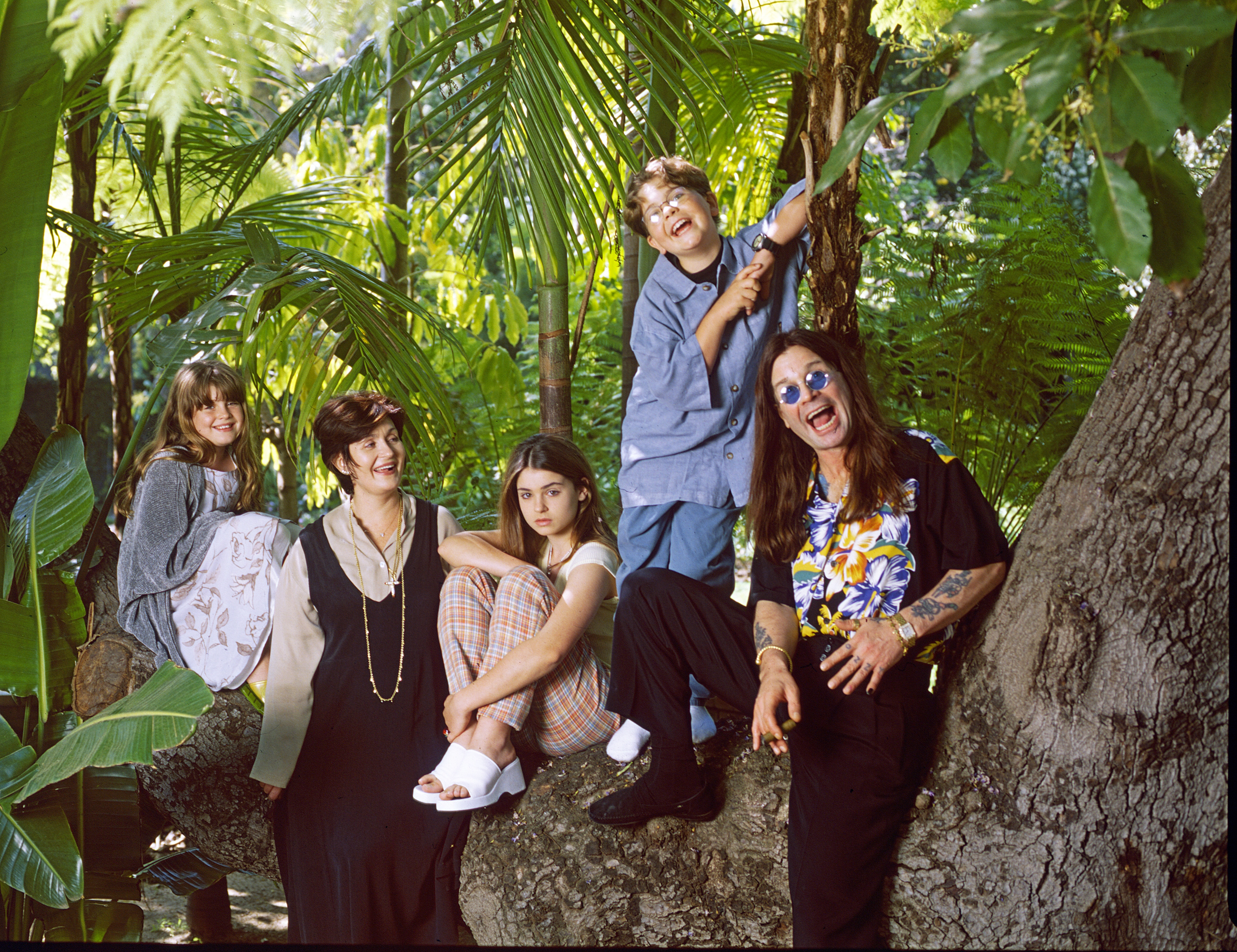 A group of five people, casually dressed, are posed outdoors among tropical plants, with one child playfully sitting on a tree branch
