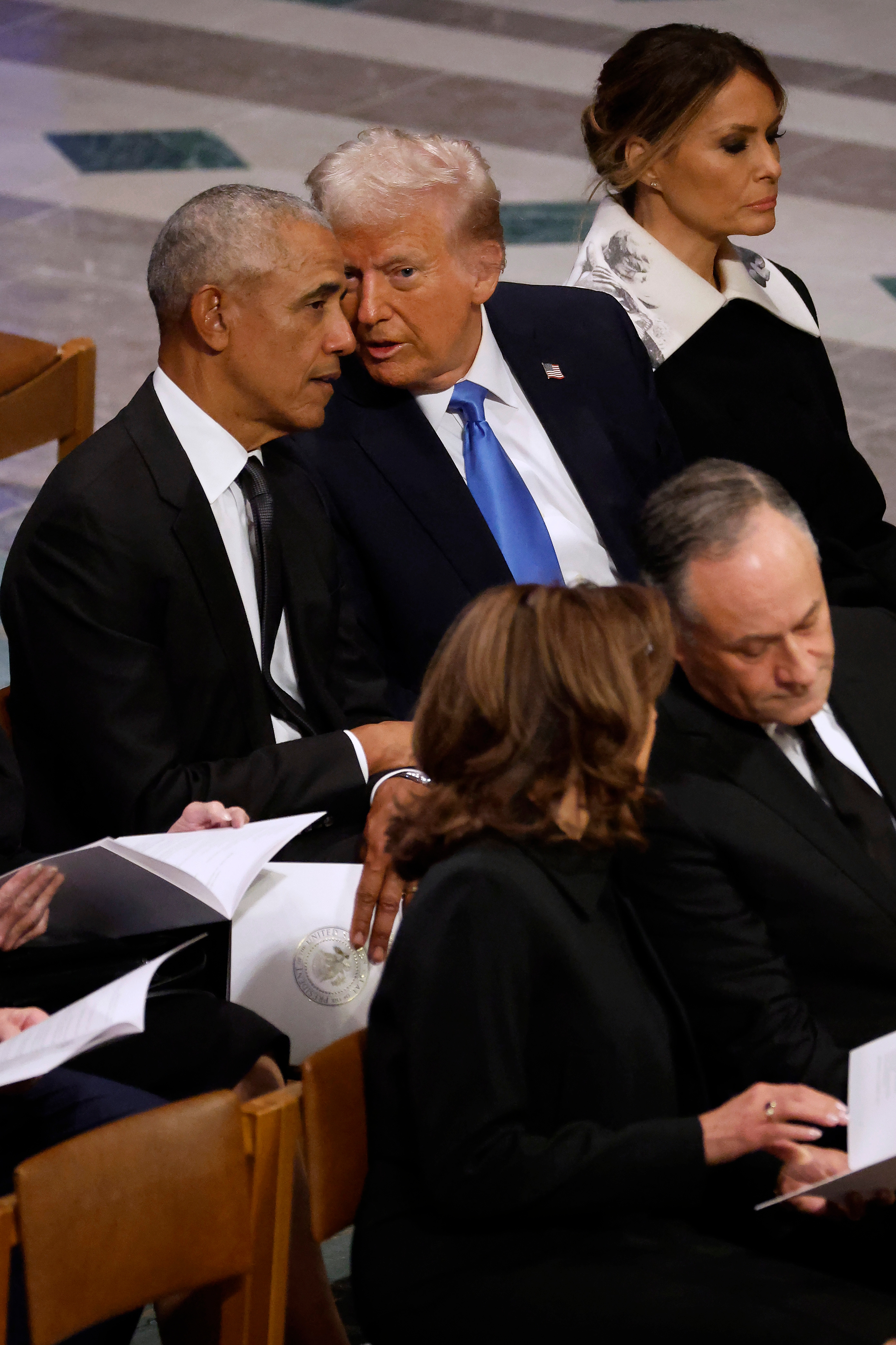 Public figures seated together at an event, two conversationally leaning in. Formal attire with suits and dresses. Event programs are in some hands