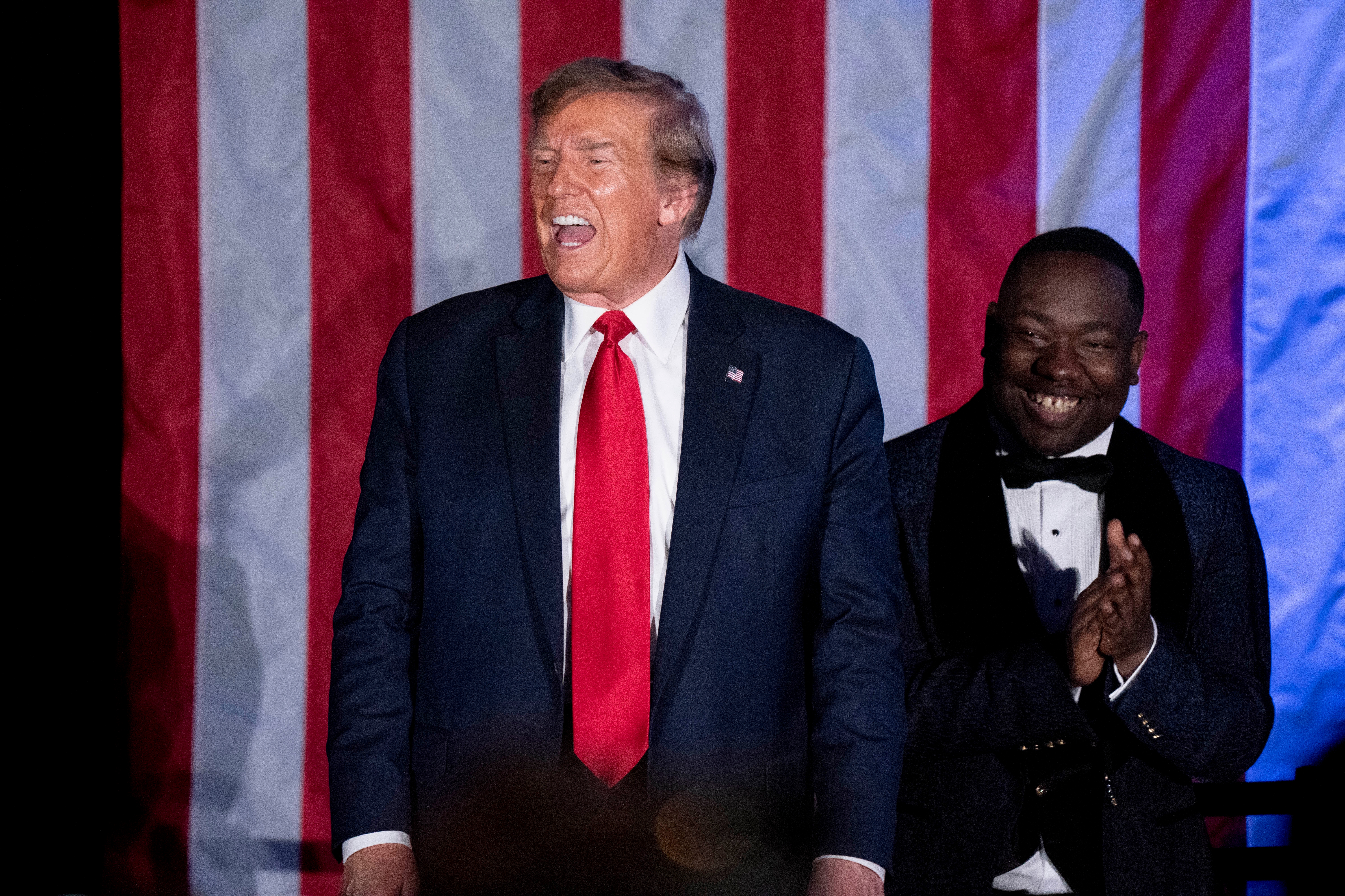 A man in a suit with a red tie stands next to another man in a tuxedo, smiling, with a U.S. flag backdrop