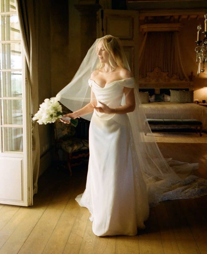 Novia con vestido de boda fuera de los hombros y velo largo, sostiene ramo de flores blancas en elegante habitación iluminada por ventana