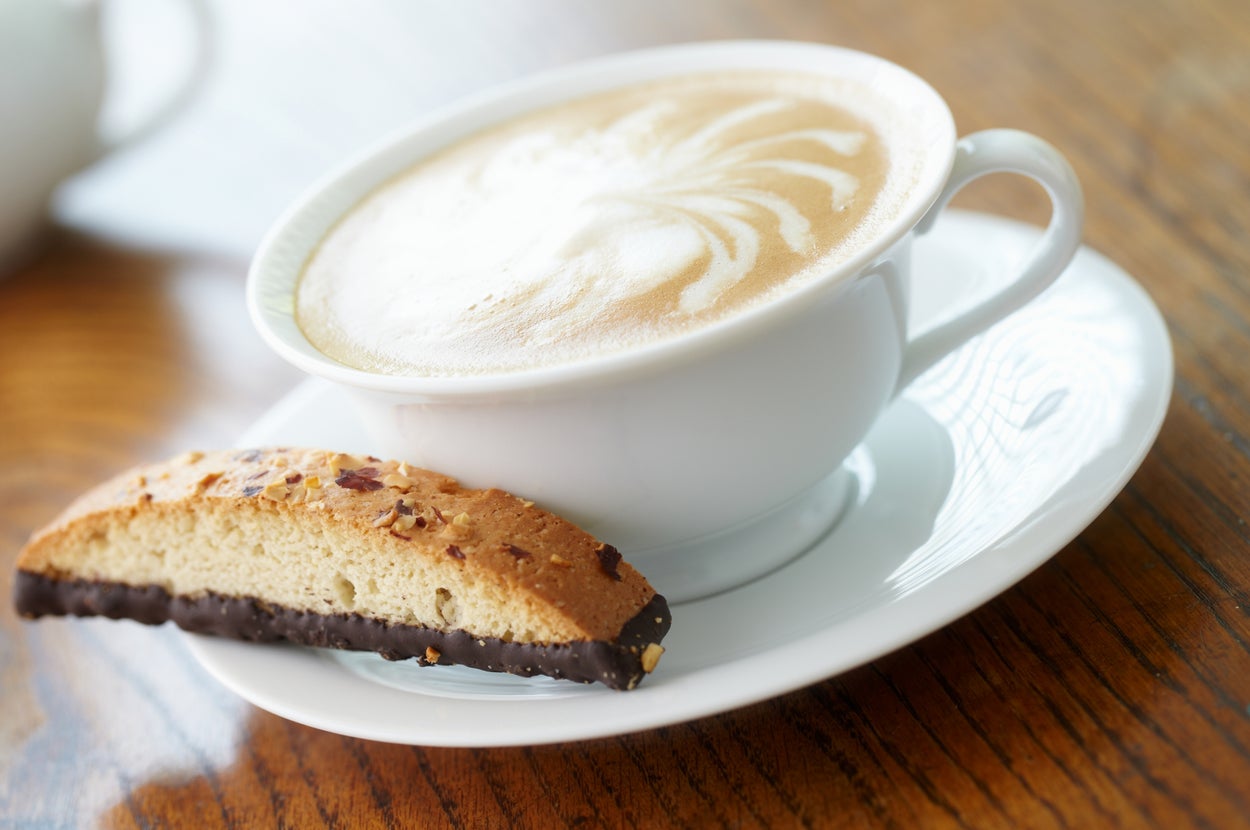 A cup of cappuccino with frothy art on top, placed on a saucer beside a chocolate-dipped biscotti on a wooden table
