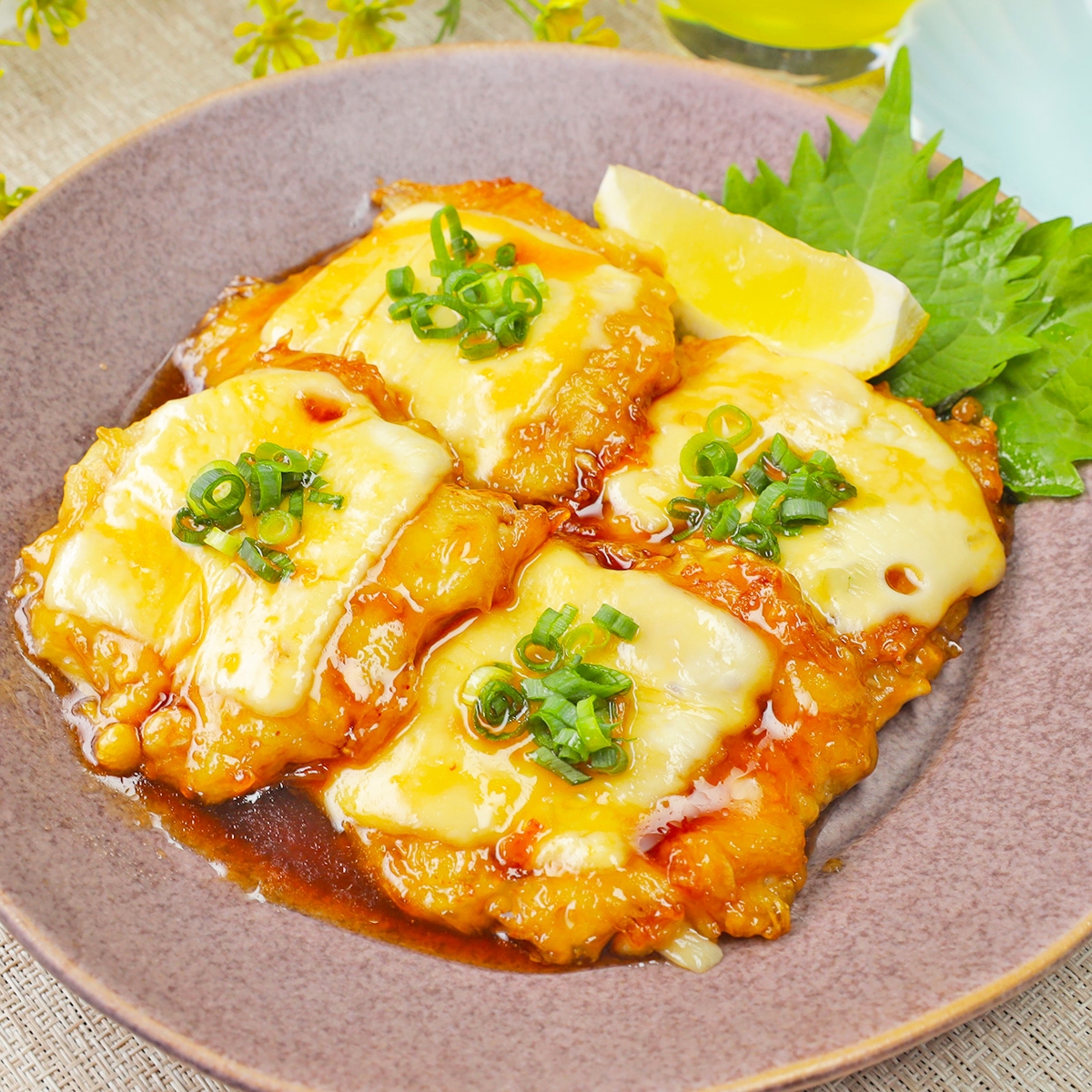 皿に盛られたチーズとネギのトッピングがされたチキンの照り焼きソースがかかった料理。レモンのくし切りが添えられている。