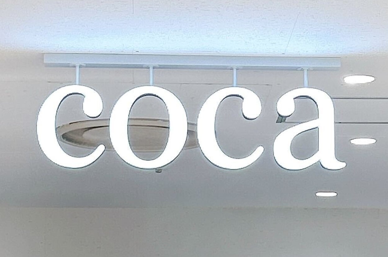店内の天井に「coca」と書かれた白いネオンサインが表示されている。背景には洋服が吊るされている。