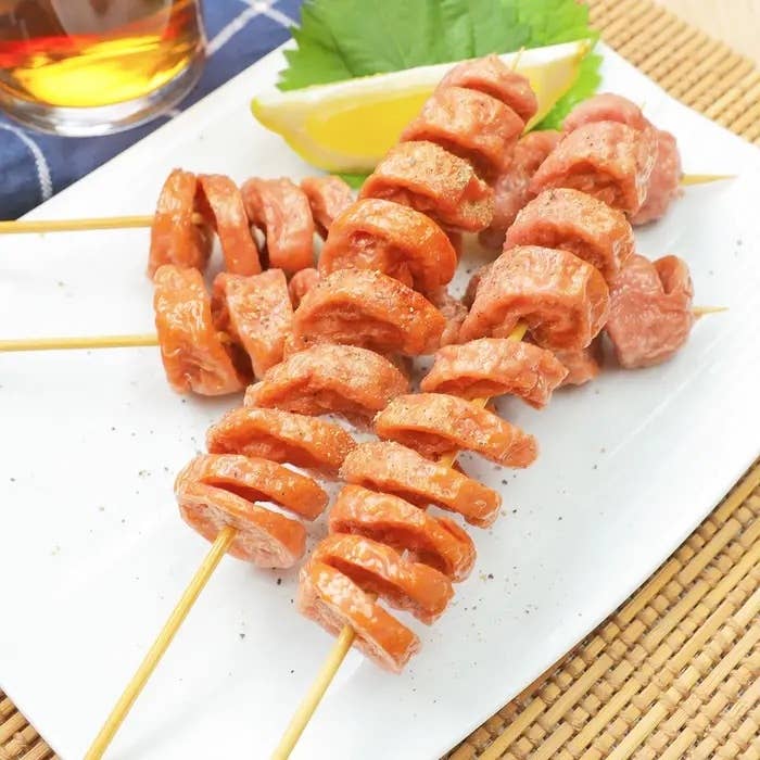 串に刺さったスライスソーセージが白い皿に盛られている。隣にレモンと青じそが添えられている。