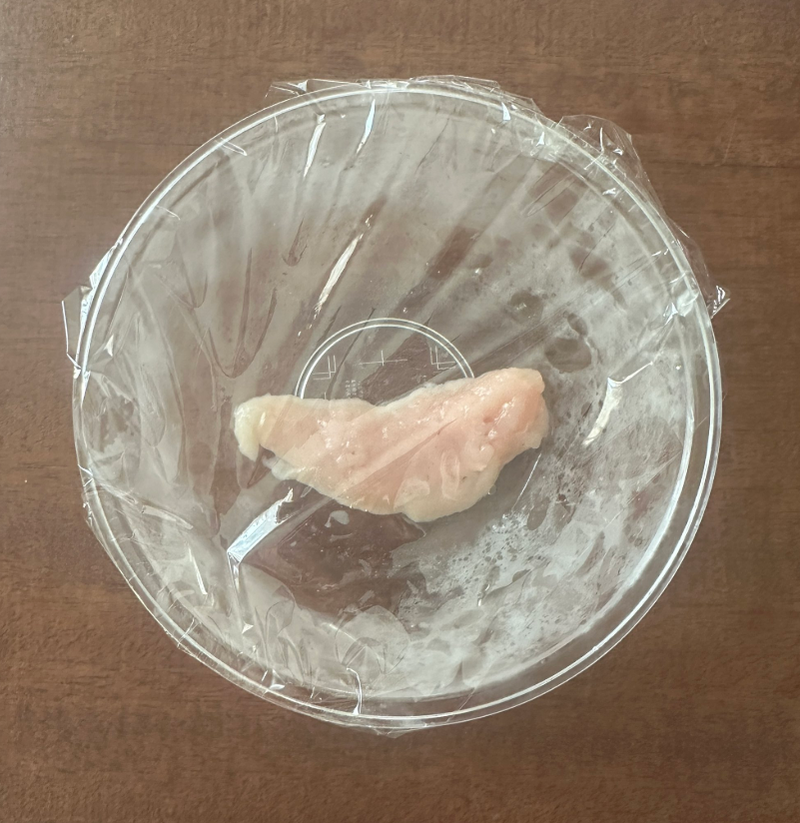 ラップで覆われた透明なボウルに入った生の鶏肉。