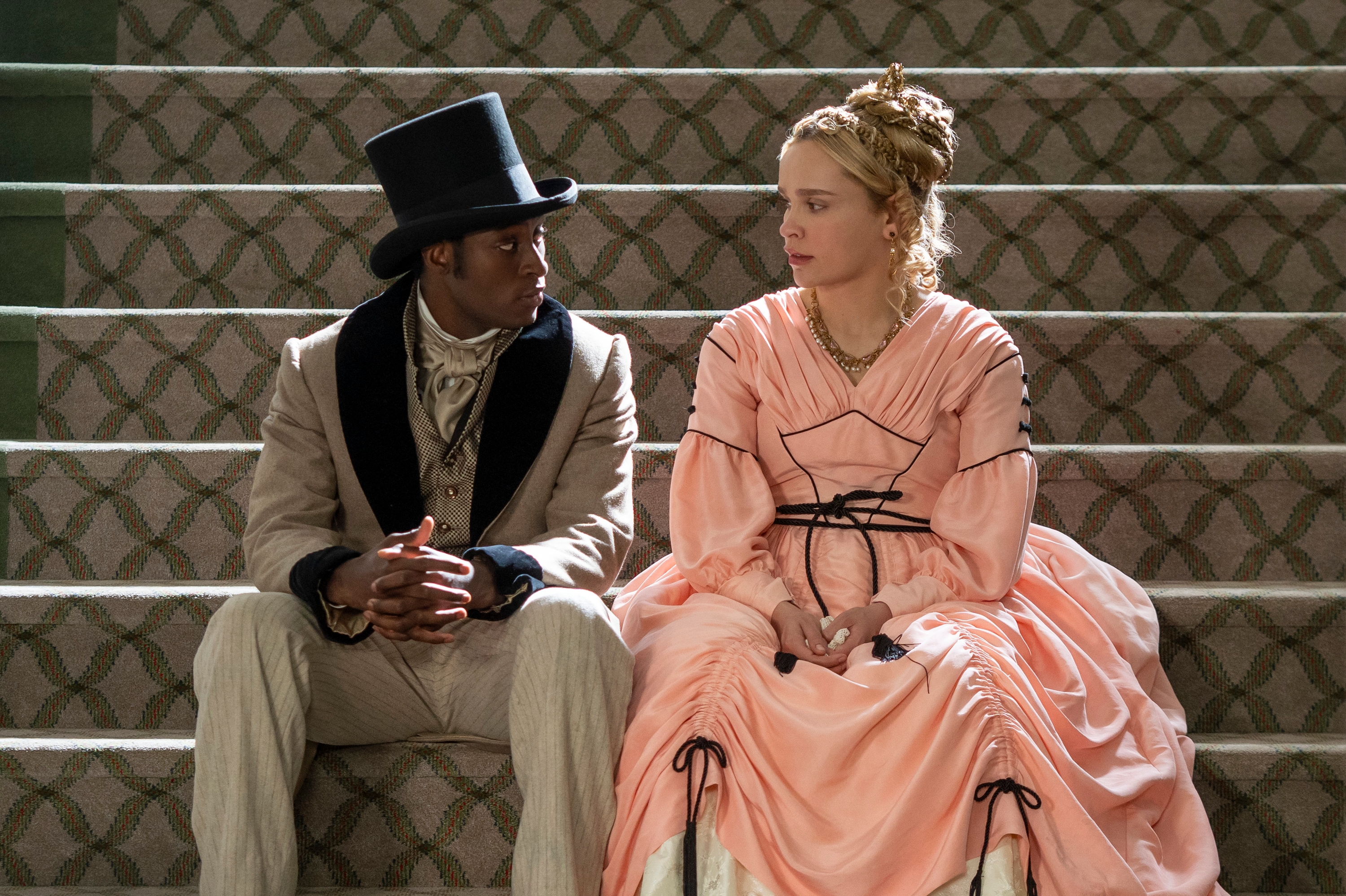Two people in period clothing sit on stairs, facing each other. One wears a suit with a top hat; the other wears a long, elaborate dress