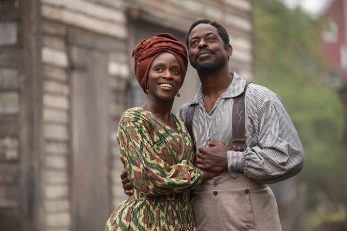 A couple poses in historical attire, with the woman in a patterned dress and headscarf, while the man wears suspenders and a striped shirt