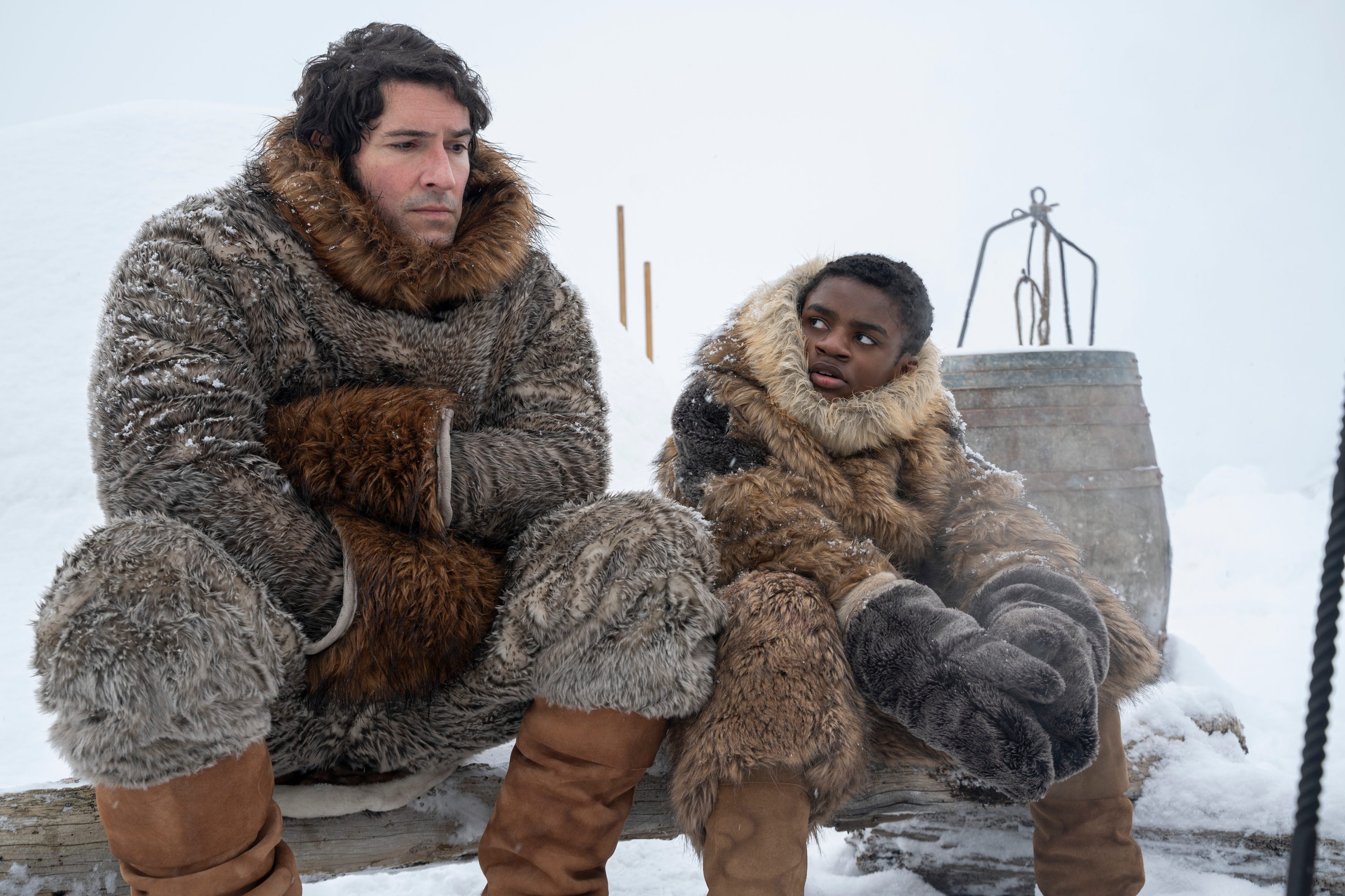 Two people in fur suits sit on a wooden bench in a snowy setting, engaging in conversation. Snowy landscape visible in the background