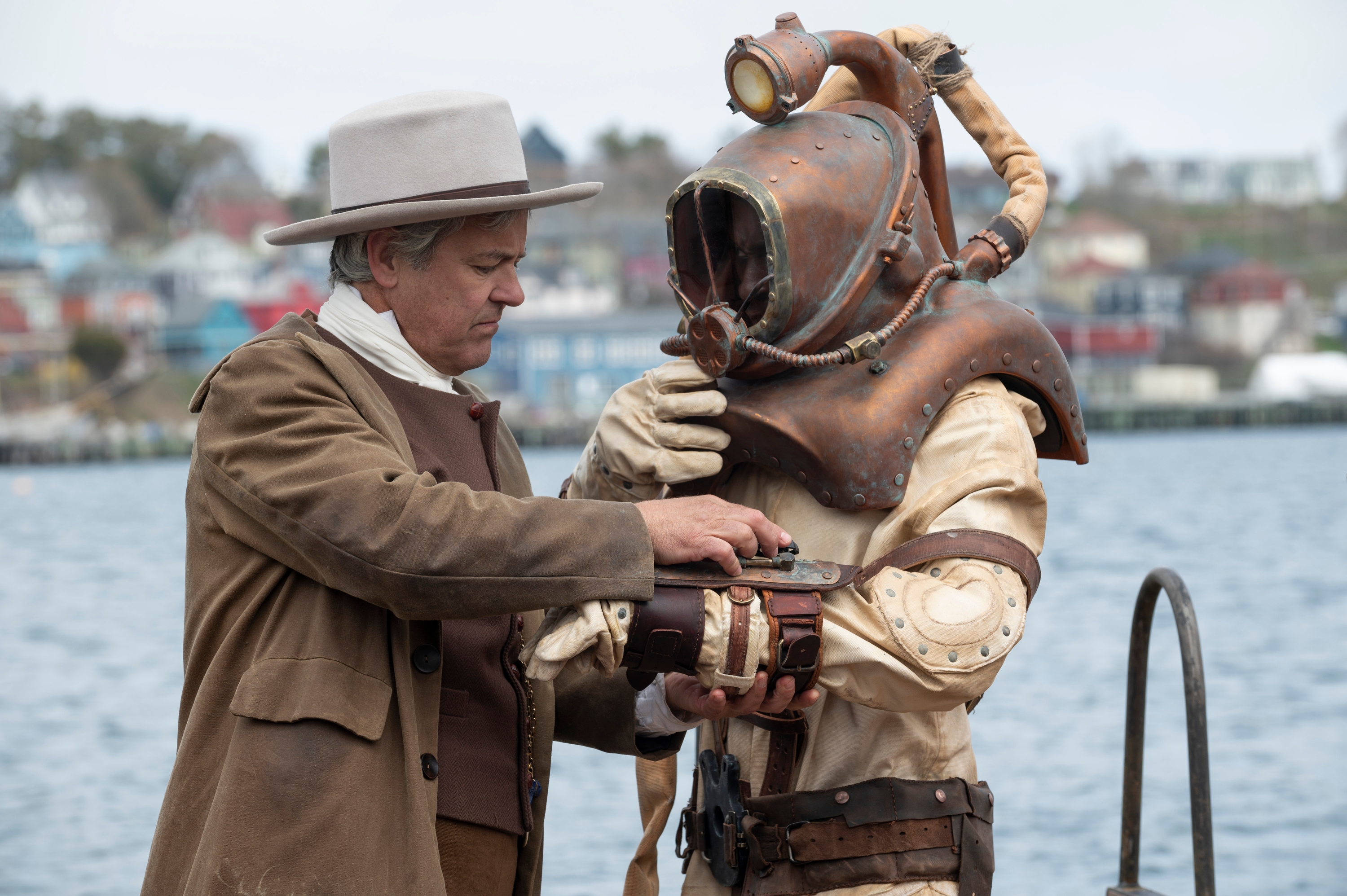 Two people near the water. One is wearing a vintage diving suit with a helmet, and the other is in period clothing adjusting the suit
