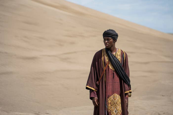 A person stands in a desert wearing traditional attire with a patterned robe and a headscarf, conveying a contemplative expression