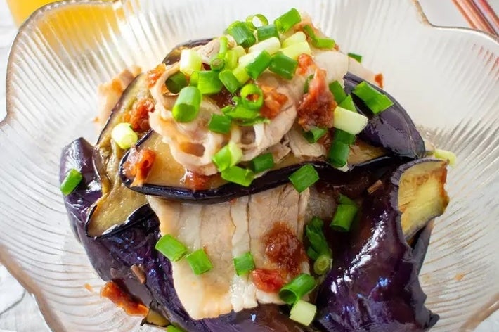 ナスと豚肉の料理が透明なガラスの器に盛られている。ねぎと調味料がトッピングされている。