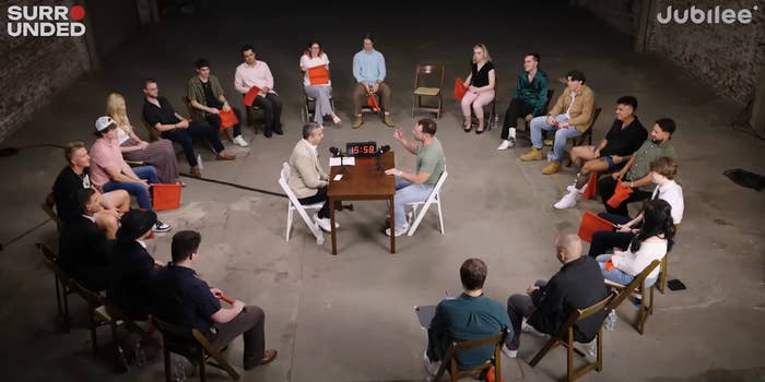 People sit in a circle for a discussion, with two men at a table in the center, participating in a recorded debate