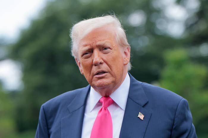 Trump in a suit and bright tie stands outdoors, with trees in the background