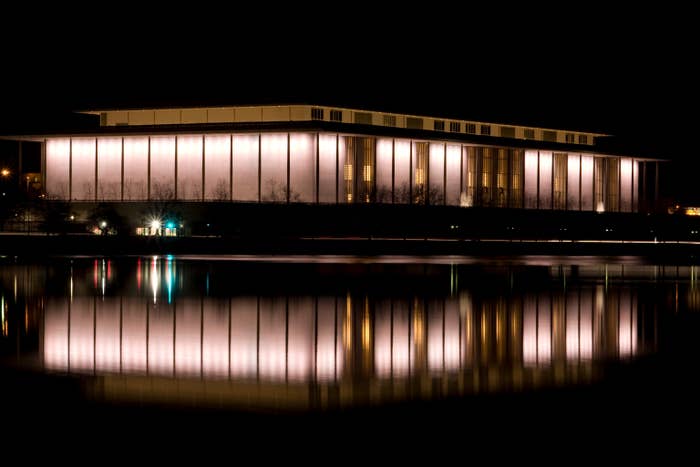 The image shows a brightly lit building at night, reflected in a calm body of water, creating a symmetrical visual effect