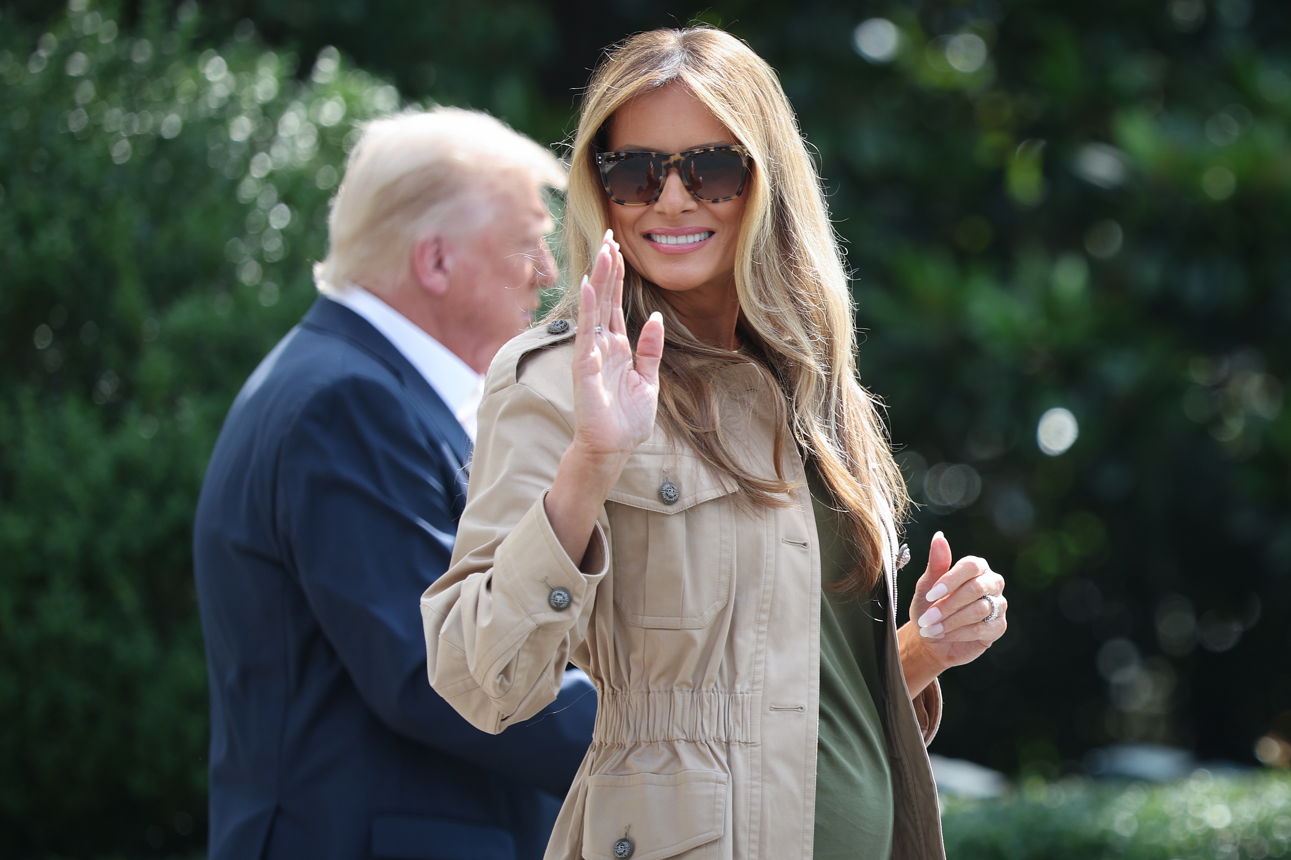 A woman in a trench coat and sunglasses waves while another person in a suit stands nearby outdoors