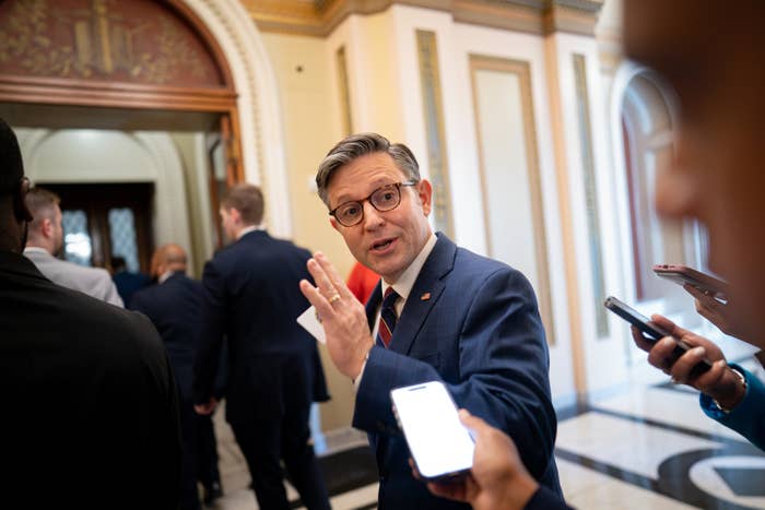 Man in a suit, holding papers, speaks to reporters with microphones in a government building hallway