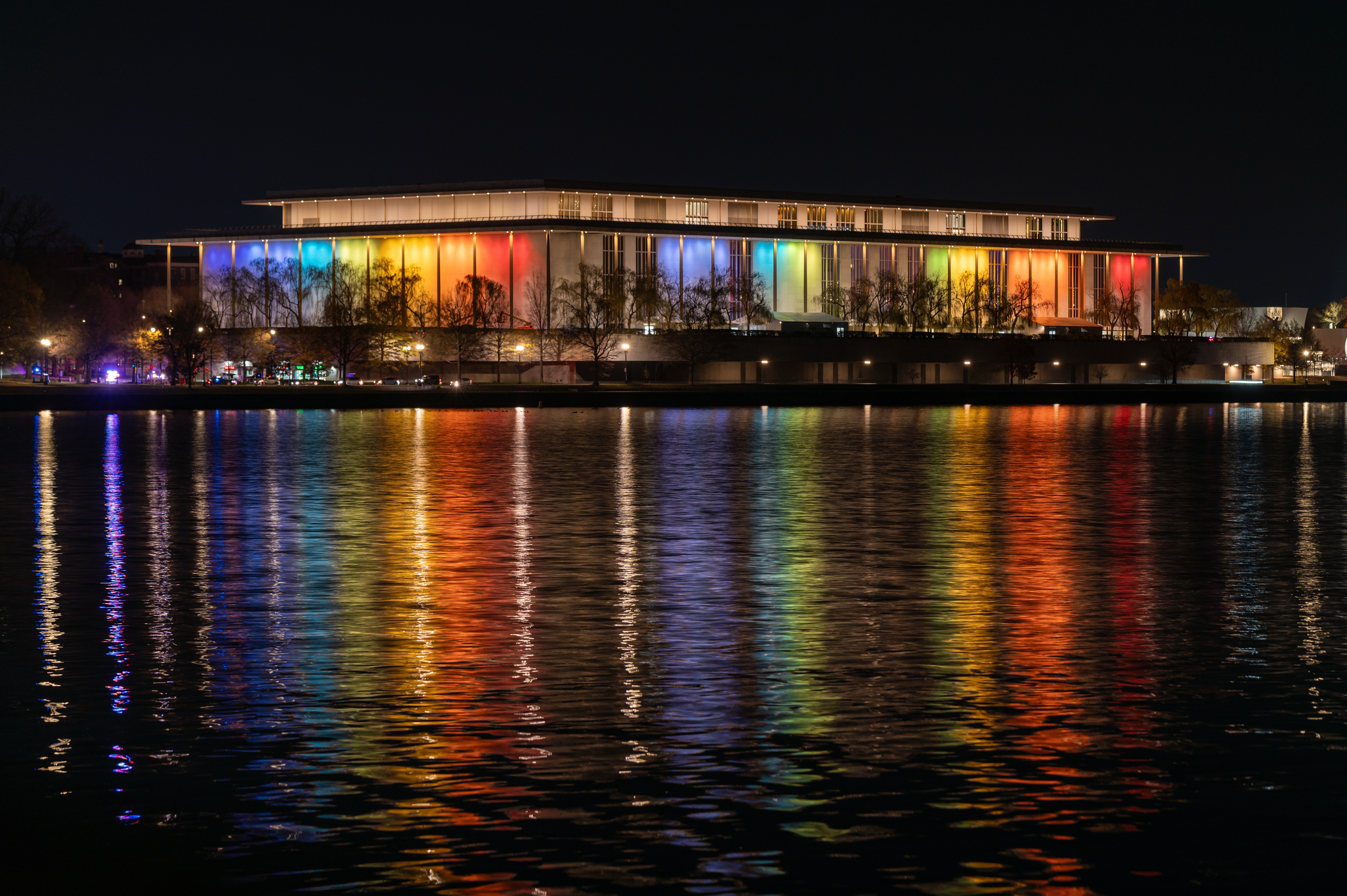 The Kennedy Center illuminated at night, reflecting vibrant lights across a calm river
