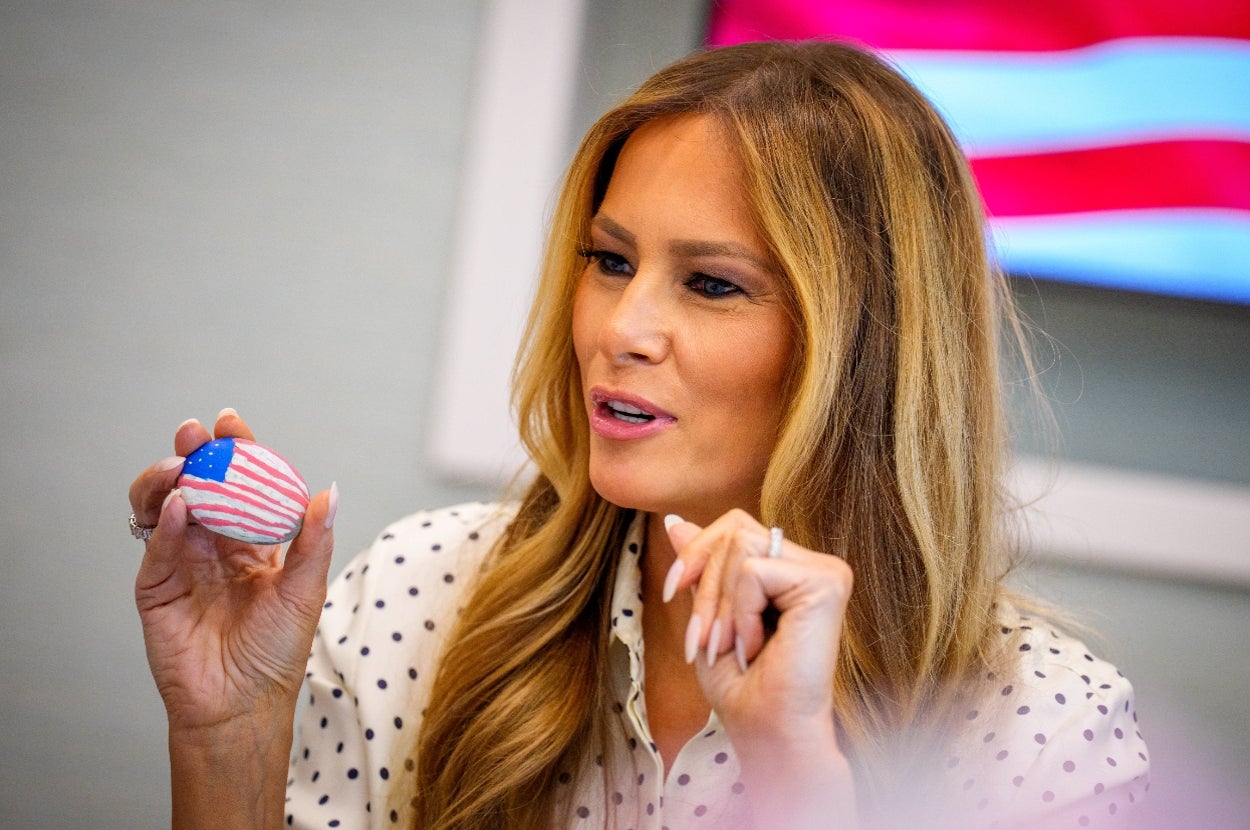 Person holding a painted egg with a U.S. flag pattern, wearing a polka dot shirt, sitting indoors with a flag visible in the background