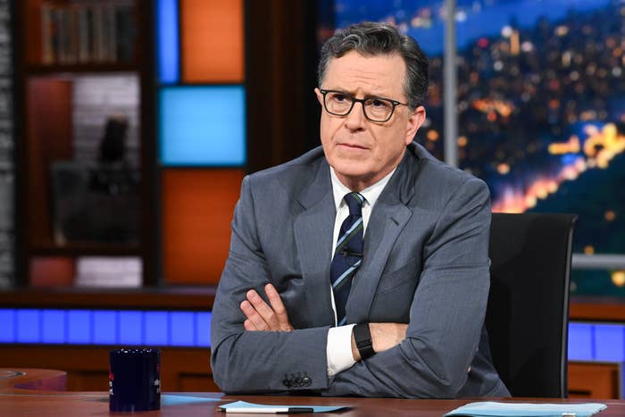 Person in a suit and tie on a talk show set, seated with arms crossed, appearing thoughtful