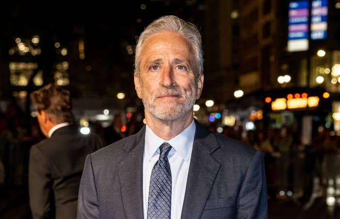 A person in a formal suit and tie stands on a busy street at night, with city lights and a crowd visible in the background