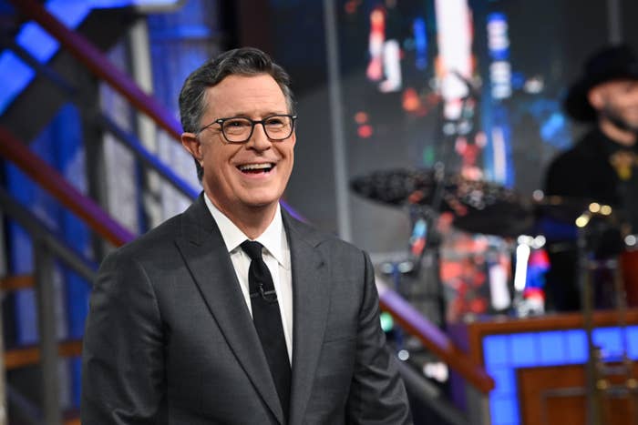 Man in a suit and tie smiles on a TV set with a band in the background