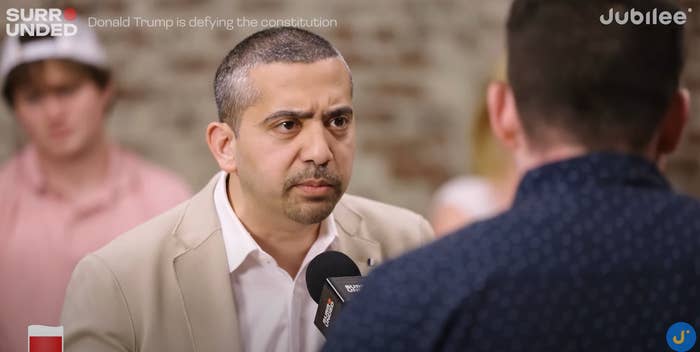 Man in a blazer holding a microphone, engaged in a serious discussion with another person