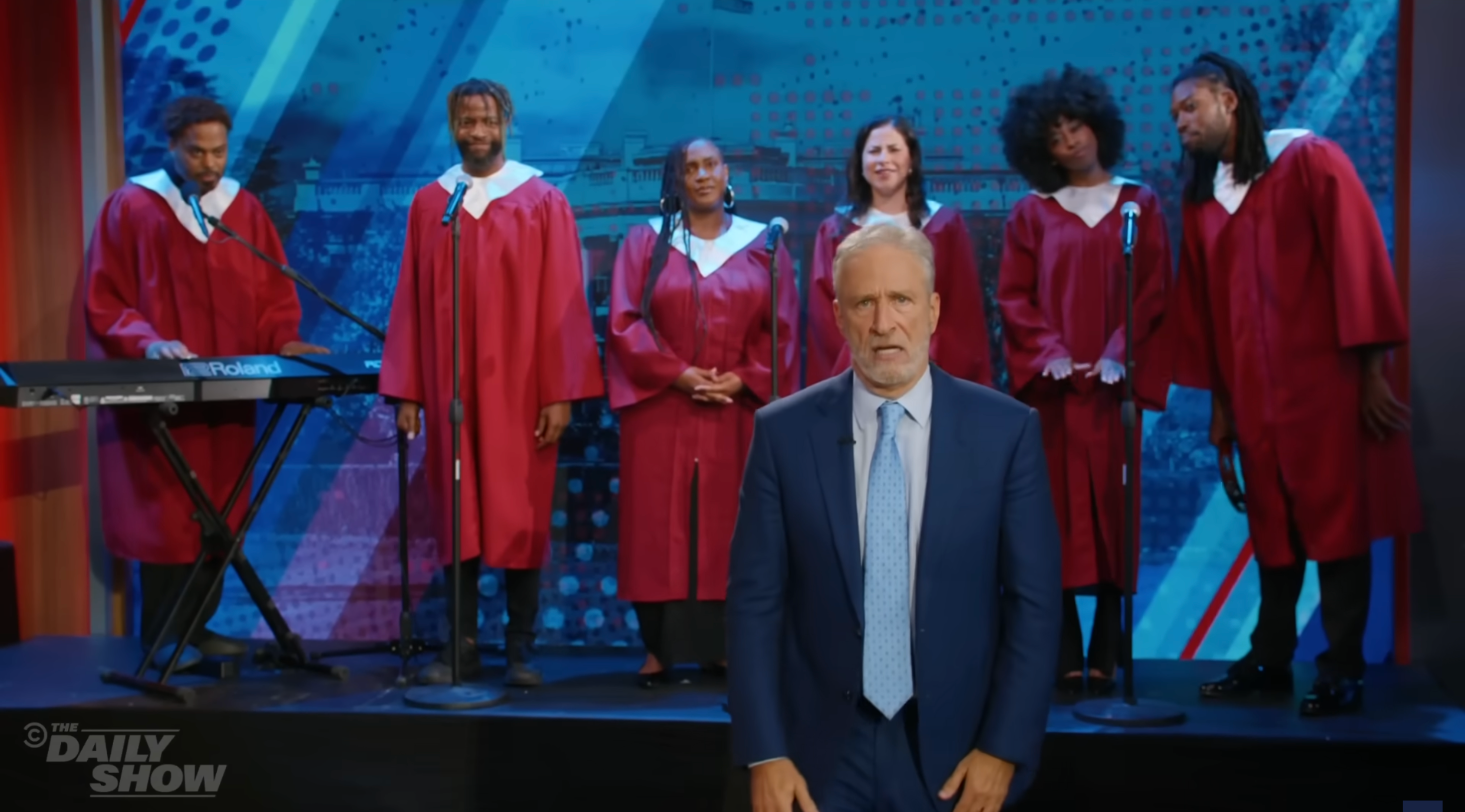 A man in a suit stands in front of a choir in red robes with a keyboard and microphones on a stage